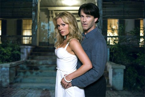 Sookie, wearing a white dress, and Bill, wearing a gray shirt and dark slacks with fangs out, are standing next to each other in front of a house.